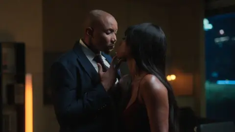 Netflix/ Iconic Digital A man in a dark suit and tie, cups a woman's chin in an intimate, close moment, their faces nearly touching in a dimly lit room with warm ambient lighting in the background.