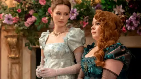 Hannah Dodd (left) as Francesca Bridgerton and Nicola Coughlan as Penelope Featherington in episode 301 of Bridgerton. The women are wearing evening dresses and standing in front of flower decorations.