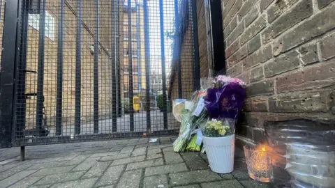 Floral tributes can be seen outside the gates of the flats, as well as a lit candle 