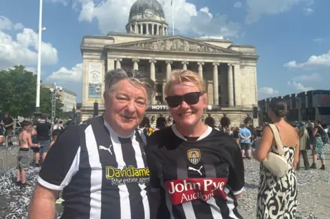 A man and a woman smiling for the camera in Nottingham's Old Market Square - they are both wearing black and white-striped Notts County shirts.