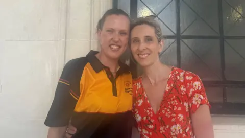 Jenna Thevanau and Candace Street. Jenna has brown hair tied back, wearing a yellow and black jersey. Candace has blonde hair tied back wearing a red floral dress. Both have their arms around each other.