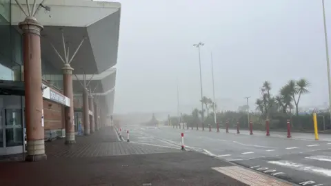 A foggy Jersey airport, with the departures building seen to the left and the road covered in mist on the right.