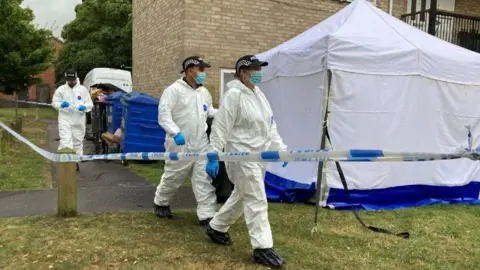 Andy Trigg/BBC Three officers in white suits walk through a police line with a white tent.