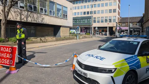 Alex Moss/BBC Police car parked in front of a closed off street, which is being guarded by a police officer behind tape.