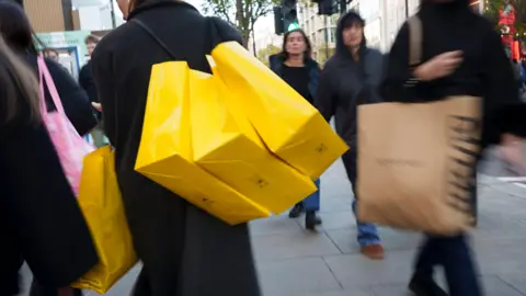 Getty Images Shoppers dressed in woollen coats and puffer jackets walk with their bright yellow Selfridges shopping bags down Oxford St in London