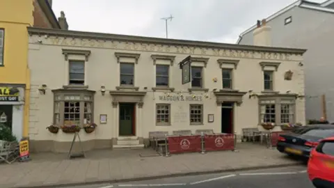Google Pub exterior showing flat roofed cream-coloured building with seating outside and a branded barrier