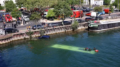 The River Seine in Juvisy-sur-Orge. The roof of a bus can be seen under the water, on which a small group of people are sitting. On the road nearby there are police cars and fire engines.