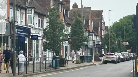 Shops line the road in West Byfleet with railings along pavements, where people are walking past the businesses. There are trees dotted along the road and further in the background. Cars are parked by the roadside and there are lampposts and road signs.