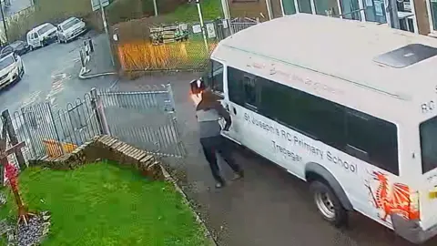 A man with his hood up stands next to the white minibus
