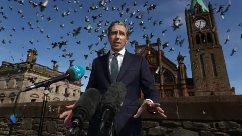 PA Media A suited Simon Harris stands on Derrys Walls with the guidlhall behind him. A flock of pigeons flies overhead.