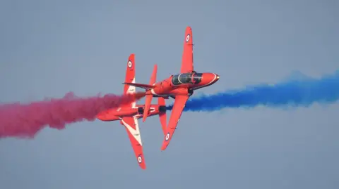Two red jets pass each other at close distance while releasing red and blue smoke behind them.