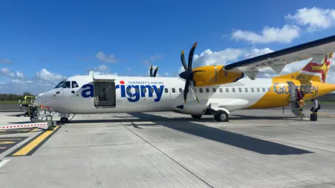 An Aurigny aircraft on a runway as two passengers board the rear of the yellow and white plane. The aircraft has twin propellers.
