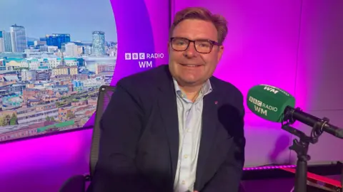 Cllr John Cotton, Labour leader of Birmingham City Council sitting in the BBC WM studio in front of purple studio lighting. He is looking into the camera smiling and leaning against the desk