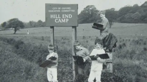 A black and white archive image of school children standing in a field next to a sign for Hill End.