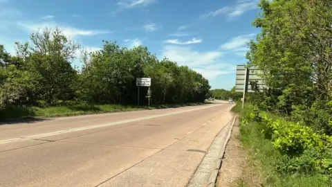 Old tarmac road. There's a duel-carriageway with trees on the side. There's a road sign on the side of the road.