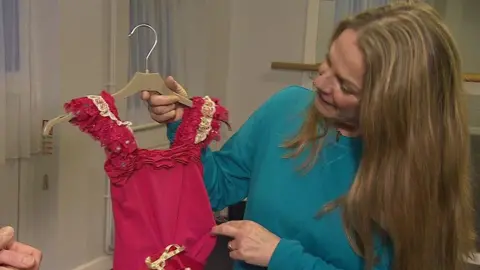 A woman with long blonde hair wearing a teal jumper holds up a child's small pink leotard with ruffled straps as she stands in a dance studio. She is turning her head to look at the leotard as she shows it off.