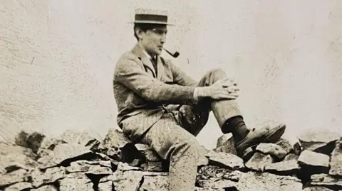 Photograph of the Cheshire mountaineer George Mallory. The black and white image shows him sitting on a dry stone wall.