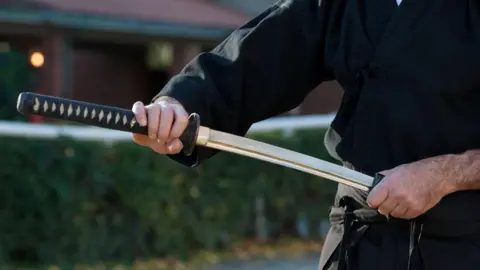 A samurai sword is brandished in an outdoor area by a man dressed in black
