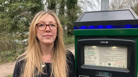 A women with brunet hair, wearing brown glasses and a black jumper. She is stood next to a green parking meter.