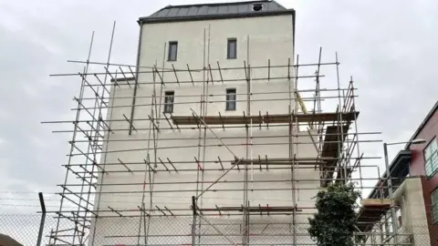 A side profile of the building. It has a white rendering and is covered in scaffolding. There are clouds above.