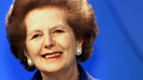 Margaret Thatcher smiling against a dark blue background. She is wearing big pearl earrings with red lipstick and has brown hair in a bouffant, swept-back style.