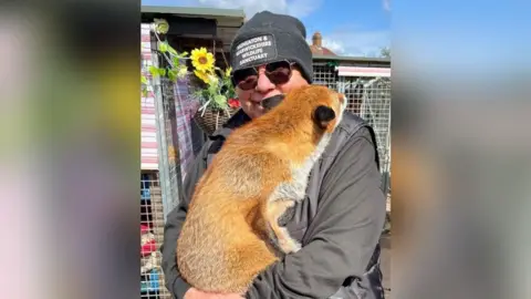 A man in a black uniform smiles at the camera while holding a fox, who leans its head on his shoulder