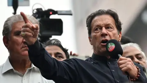 Imran Khan wearing a black button-up shirt and speaking into a microphone, points dramatically. Behind him are other men, and a television camera