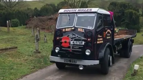 Family photo A old black lorry with a white roof driving down a narrow path with grass on both sides and gravestones on the left hand side