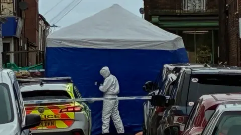 A street with multiple parked vehicles, including a police car, and a blue and white forensic tent. A person in a white forensic suit stands near the tent inside the police cordon.