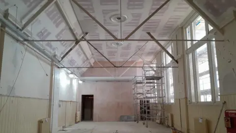 Ipswich Museums View of a room with a high ceiling which is painted pink and white. The walls of the room are yellow and white, and there are two scaffold towers, made of silver metal, against the right wall. 