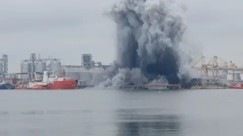 A large cloud of dark smoke is seen from a distance rising from a port. Boats, cranes and silos can be seen around it.
