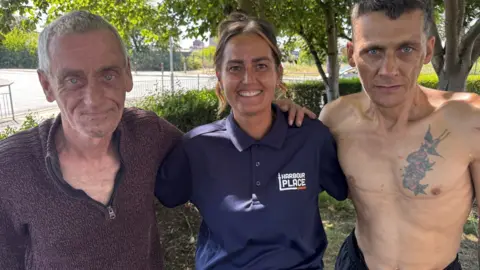 Crispin Rolfe / BBC Stacy Brown who has brown hair and is wearing a blue polo shirt with the Harbour Place logo on stands between two men at the support centre