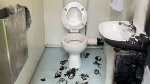 Redcar and Cleveland Borough Council The single toilet cubicle has been vandalised. The sink on the right is blackened and torched from fire damage. There are burn marks on the blue floor and the white wall above the toilet is blackened also. There is a metal bin on the wall to the left. 