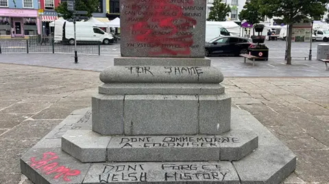 The David Lloyd George statue with black graffiti with the words 'for shame', 'don't commemorate a coloniser' and 'don't forget Welsh history' daubed on the plinth. The town high street is in the background with traffic and shops visible.