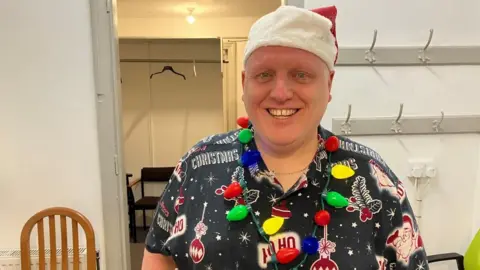 BBC/Seb Cheer A man wearing a shirt with "Christmas" and "Ho Ho" written alongside drawings of Santa, wearing a light-up garland around his neck and a Santa hat. He is standing in a theatre dressing room with wardrobe rails and coat hooks behind him.