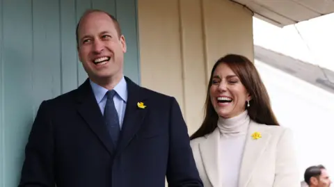 Getty Images Prince William and Catherine standing side by side. William is standing on the left wearing a black suit and blue shirt with a daffodil pin on his lapel. Catherine is standing on the right wearing a cream blazer and a white turtle neck top, she also has a daffodil pin on her blazer. She has pearl drop earrings. Both are laughing. 