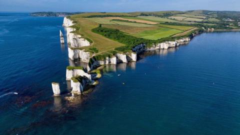 TUESDAY - An aerial photograph of the white cliffs at Old Harry Rocks on the Dorset coast. There are several stacks of cliff on this stretch of coast that stick out into the sea. Behind there are green frields and in the foreground the sea is a bright blue on a sunny day