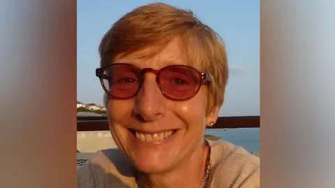 Family photograph Ruth Hayhurst with short light brown hair wearing sun glasses smiling with sea in the background