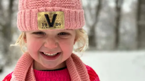 Alan Johnstone A close-up photo of six-year-old Sienna smiling to camera wearing a pink hat and scarf and a pink jumper with a snowy scene in the background