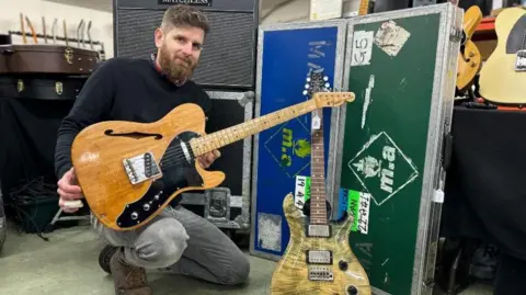 Gardiner Houlgate Auctioneers Auctioneer Luke Hobbs on one knee holding one of the guitars which is on sale. He is wearing grey jeans and a black jumper. Another of the guitars is on a stand.