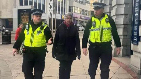 A woman with grey hair tied back in a black coat and black trousers is walking through Sheffield city centre with two male police officers either side of her