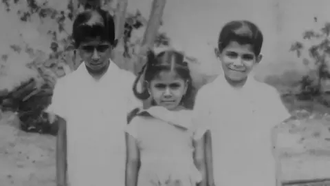 Tara Mistry A black and white photo showing a young Tara Mistry standing in the middle of two boys. The boys are wearing white shirts while Tara wears a white dress.