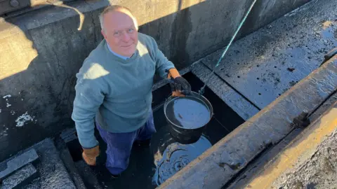 A man in a light blue top and blue trousers is looking up at the camera, holding a bucket and standing in water