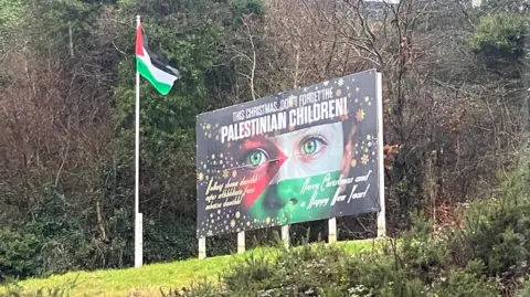 The picture shows a billboard up on a grassy hill saying "This Christmas don't forget Palestinian children" with a Palestinian flag beside it
