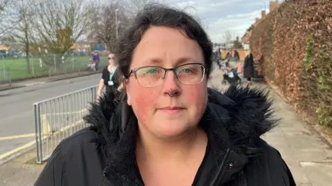 John Devine/BBC Wendy Fieldhouse is standing on a pavement next to a road. She has dark brown hair in a ponytail and is wearing black-framed glasses and a black coat. 