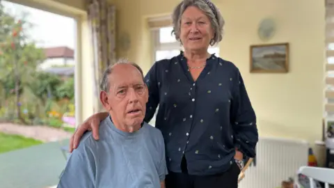 Lynne Telford stands with her husband Rick, who lives with Parkinson's disease, who is sitting down in their home.