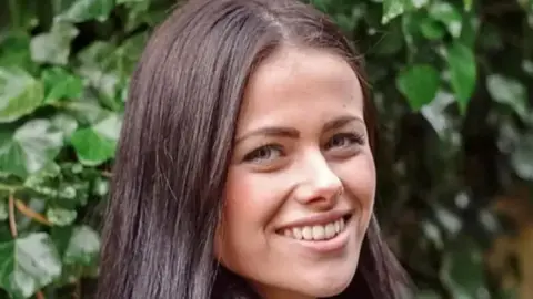India Chipchase, a young woman who is looking directly at the camera and smiling. She is facing towards the right and has long dark straight brunette hair. She is standing outside infront of a green leafy bush.