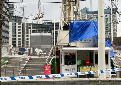 A shot of the ride in daylight, showing white plastic seats at an angle with chains twisted together, and the entrance to the ride in the foreground that's been covered up with tarp. The area is cordoned off with police tape.