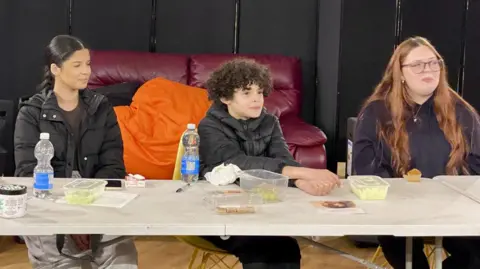 A teenage girl with mixed heritage hair, another teenage girl with long red hair and an 11-year-old boy with afro hair are smiling while sitting at a table during a workshop. They have plastic boxes with green coloured hair care masks. 