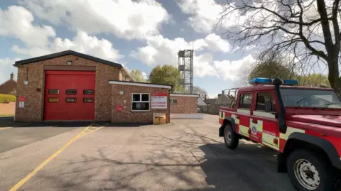 The forecourt of a modestly-sized single-storey fire station with a large single red garage door. A Land Rover-type vehicle in fire service livery is parked outside.